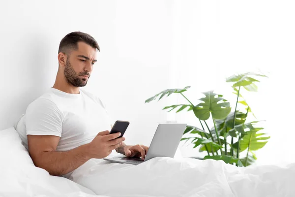 Счастливый кавказский привлекательный молодой парень в белой футболке печатает блог на компьютере и смотрит в телефон — стоковое фото