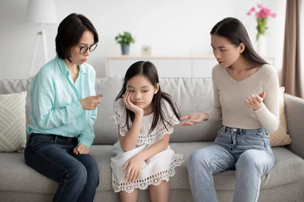 Asian mother and grandmother scolding unhappy little girl on sofa at home