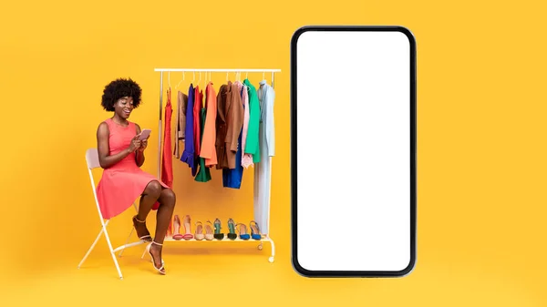 Africano femmina seduta vicino a grande telefono shopping su sfondo giallo — Foto Stock