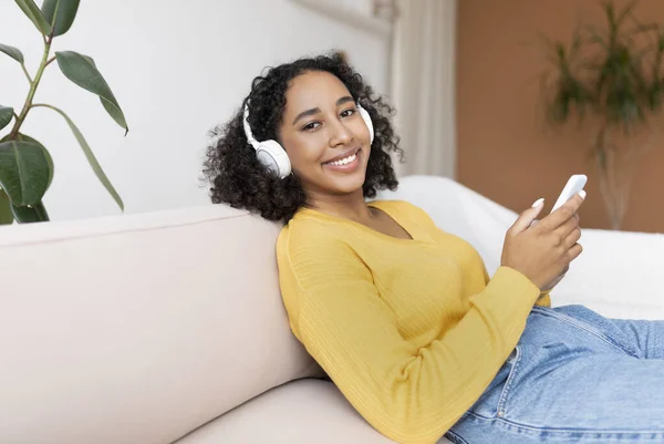 Giovane donna nera in cuffia rilassarsi sul divano, ascoltare musica o guardare film sul cellulare a casa — Foto Stock