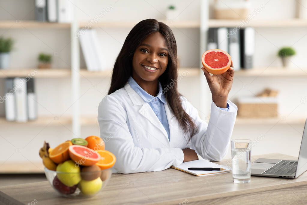 Friendly black female dietitian holding grapefruit and smiling at camera, offering online weight loss consultations