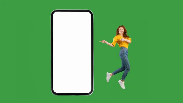 Молодая леди указывает на пустой экран смартфона прыжки, зеленый фон — стоковое фото