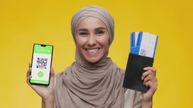 Orta Doğulu genç mutlu bir kadın uçuş biletleri ve akıllı telefon uygulamasında Covid-19 aşı sertifikası gösteriyor.