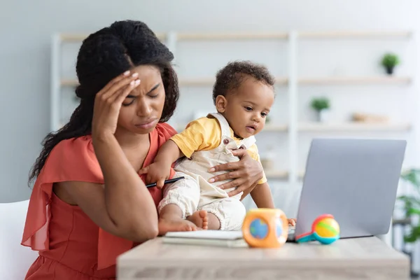 Внештатный стресс. Измученная черная женщина работает над ноутбуком с ребенком в руках — стоковое фото
