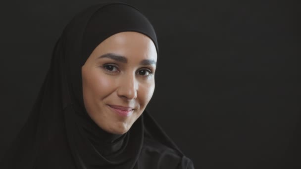 Jovem mulher positiva do Oriente Médio em lenço de cabeça tradicional sorrindo para a câmera, fundo do estúdio preto com espaço vazio — Vídeo de Stock