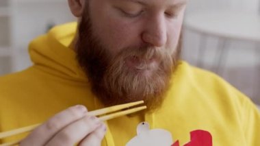 Sakallı adam erişte yiyor. Yemek çubukları kullanıyor. Kapalı mekanda yemek yiyor.