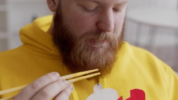 Hombre barbudo comiendo fideos usando palillos almorzando en interiores, de cerca — Vídeos de Stock
