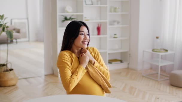 期待の概念。幸せな妊娠中のアジアの女性は赤ちゃんの体のスーツを保持し、腹に触れる — ストック動画