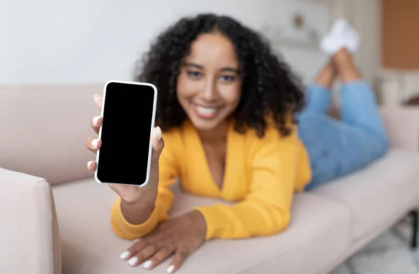 Donna afroamericana che mostra smartphone con schermo bianco, sdraiata sul divano di casa, offrendo mockup per app mobile — Foto Stock