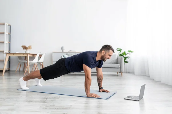 Happy millennial muscular caucasian male doing plank and looking at computer watch online lesson in living room — Stock Photo, Image