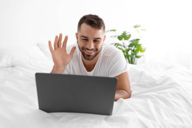 Gülümseyen yakışıklı milenyum kaslı beyaz adam bilgisayarda yazı yazıyor, dizüstü bilgisayarında el sallıyor web kamerasıyla yatakta yatıyor.