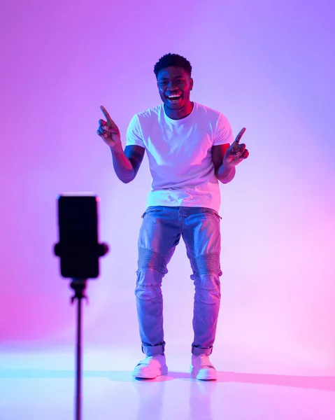 Młody Afroamerykanin męski influencer nagrywa zawartość wideo, uśmiecha się do smartfona kamera internetowa w neonowym świetle — Zdjęcie stockowe