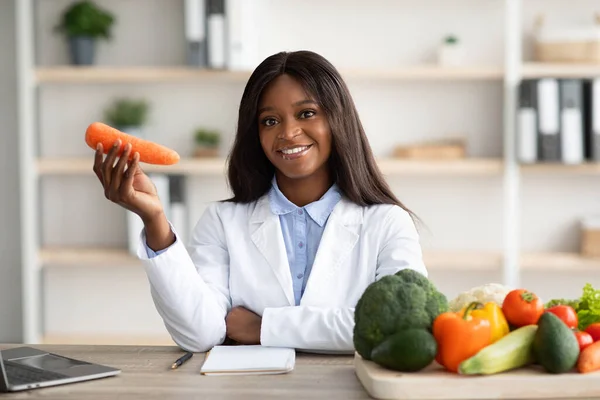 Vidám afro-amerikai női dietetikus, aki répát tart az orvosi klinikán, és laptoppal ül a munkahelyén. — Stock Fotó