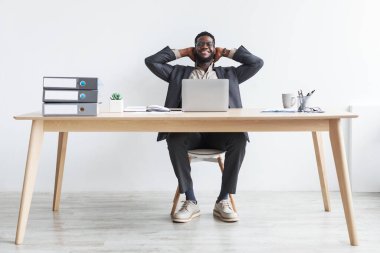 Rahat genç siyah iş adamı ofis masasında oturuyor ve ellerini başının arkasına koyup dizüstü bilgisayar kullanıyor.