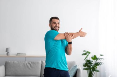 Neşeli milenyum kaslı çekici Avrupalı erkek sporcu kol egzersizi yapıyor, oturma odasında ısınıyor.