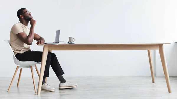 Pensiv ung svart kille som använder laptop, sitter vid skrivbordet, tänker över något, arbetar online från hemmakontoret — Stockfoto