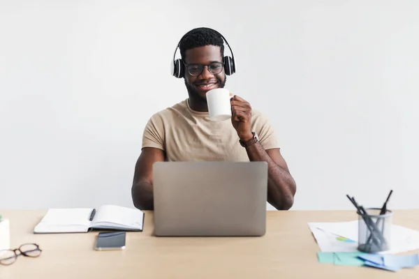 Веселий молодий чорний хлопець в навушниках п'є каву, спілкується онлайн на ноутбуці за столом на білому тлі — стокове фото