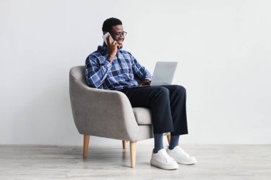 Dizüstü bilgisayarı olan, cep telefonuyla iletişim kuran, koltukta oturan neşeli, Afro-Amerikan genç adam.