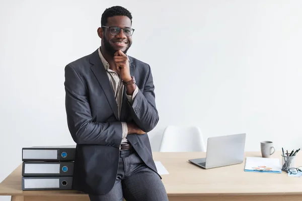Портрет молодого чорного бізнесмена в офіційному одязі і окулярах, посміхаючись на камеру, сидить на робочому столі в офісі — стокове фото