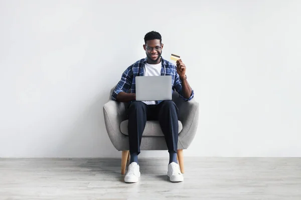 Bonito jovem negro com cartão de crédito e laptop sentado em poltrona, comprando bens na internet. Compras online — Fotografia de Stock