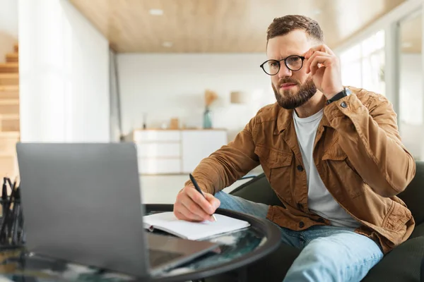 Geschäftsmann blickt auf Laptop mit Brille und macht sich Notizen zu Hause — Stockfoto