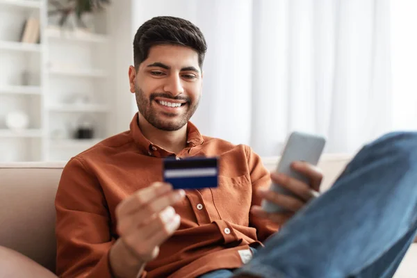 Sonriente chico árabe usando el teléfono y la tarjeta de crédito en casa — Foto de Stock