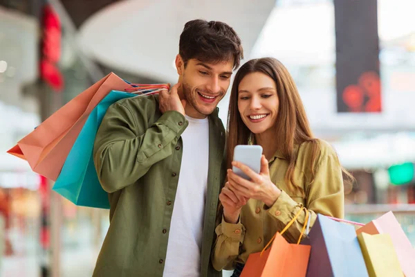Compras de casal usando aplicativo de telefone segurando sacos de Shopper no shopping — Fotografia de Stock