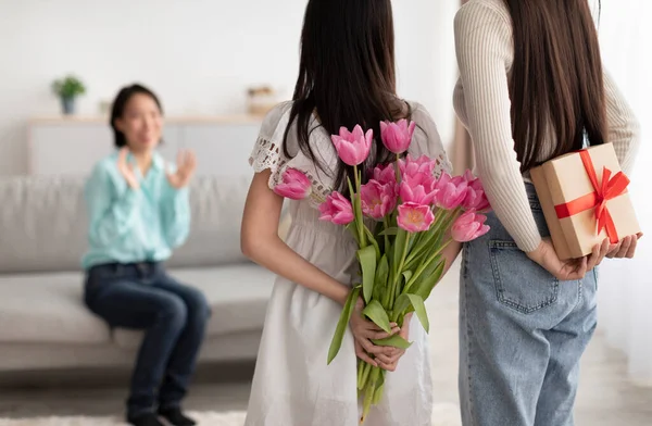 Азиатка с мамочкой, прячущей тюльпаны и подарочную коробку для пожилой женщины за их спинами, здоровается с весенним праздником дома — стоковое фото
