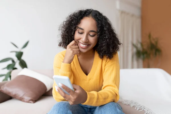 Jovem negra usando celular, tendo videochamada online, sentada no sofá, se comunicando em rede social em casa — Fotografia de Stock