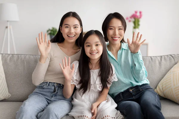 รูปภาพของเด็กสาวเอเชียที่มีความสุขกับแม่และยายนั่งบนโซฟาและโบกมือที่กล้องในร่ม — ภาพถ่ายสต็อก
