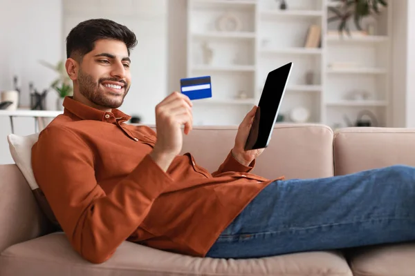 Sonriente chico árabe usando tableta y tarjeta de crédito en casa — Foto de Stock