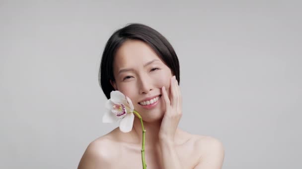 Середньолітня азіатська леді тримає орхідеї дотик до обличчя, сіре тло — стокове відео