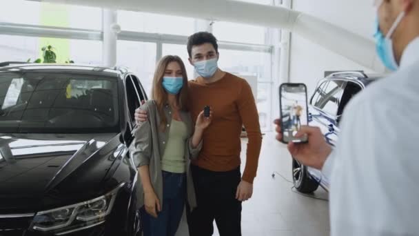 Молодая счастливая влюбленная пара в масках для лица, позирующая с новой машиной и ключами в выставочном зале. продавец фотографирует их — стоковое видео