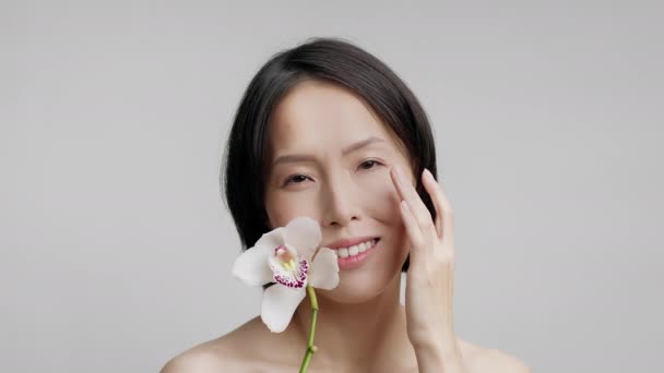 Madura japonesa mujer aplicando humectante celebración orquídea flor, fondo gris — Vídeos de Stock