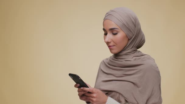 Ung vacker Mellanösternkvinna bär hijab surfing på smartphone, tittar på kameran och ler, fritt utrymme — Stockvideo
