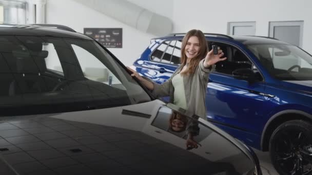 Jovem mulher bem sucedida comprando carro novo no salão de concessionárias, posando com chaves perto de novo veículo próprio, tiro de rastreamento — Vídeo de Stock