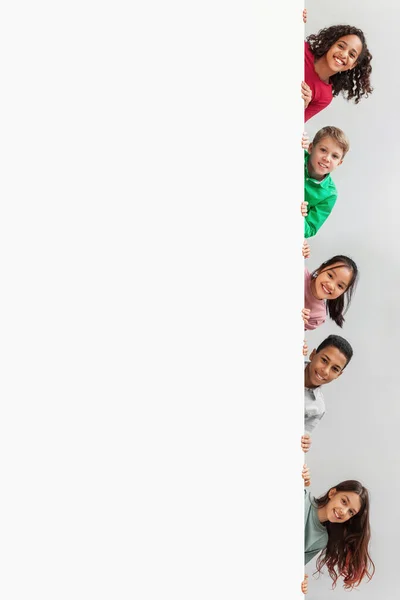 Niños multiétnicos escondidos detrás de la pizarra blanca sonriendo sobre fondo gris — Foto de Stock