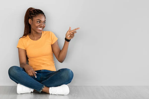 Publicidade fixe. Jovem afro-americana positiva sentada no chão, apontando para um espaço vazio contra a parede cinzenta — Fotografia de Stock