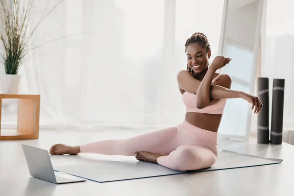 Ajuste joven mujer afroamericana estirando su brazo en casa, utilizando el ordenador portátil, viendo videos deportivos en línea —  Fotos de Stock