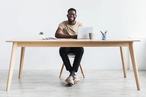 Gündelik giysiler içinde mutlu bir Afrikalı genç adam ofis masasında dizüstü bilgisayarla çalışıyor. — Stok fotoğraf
