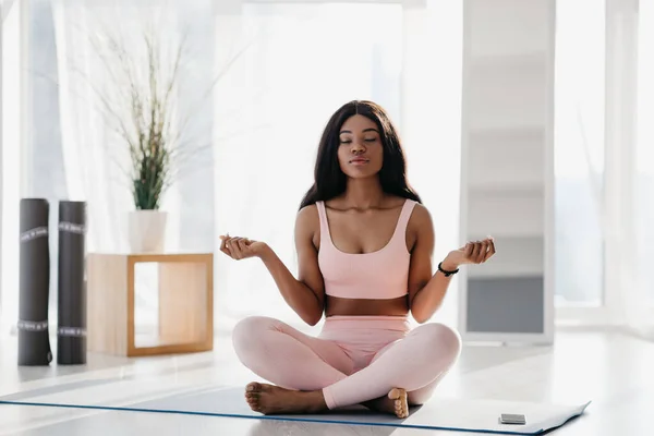 Hermosa joven negra pacífica sentada en pose de loto, meditando con los ojos cerrados, practicando yoga en la esterilla en casa —  Fotos de Stock