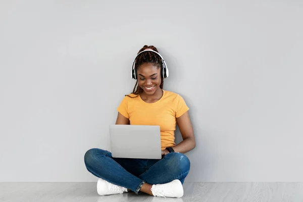Позитивна молода чорна жінка в навушниках працює на ноутбуці, сидячи хрест ногами на підлозі проти сірої стіни студії — стокове фото