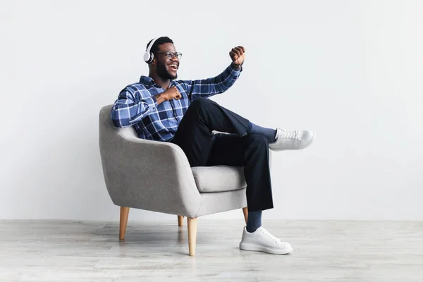 Joven chico negro alegre en auriculares inalámbricos sentado en sillón, escuchando música, bailando con su canción favorita — Foto de Stock
