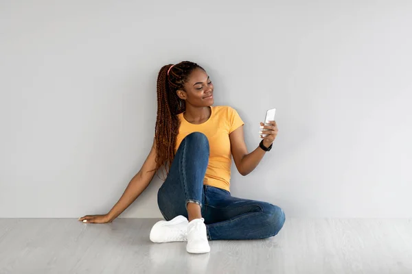 Fiatal afro-amerikai hölgy ül a mobiltelefon, böngészés internet, csevegés online ellen szürke stúdió fal — Stock Fotó