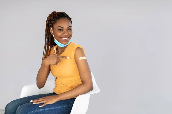Afroamerikanerin lächelt und zeigt auf Bandenhilfe, nachdem sie Coronavirus-Impfstoff auf grauem Hintergrund erhalten hat — Stockfoto