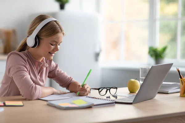 Feliz chica caucásica adolescente rubia en los auriculares toma notas, estudio en casa en la mesa con el ordenador portátil en la cocina — Foto de Stock