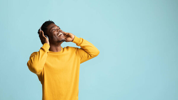 Emotional african american millennial man with wireless headset looking at copy space for advertisement and smiling over blue studio background, black guy listening to nice music