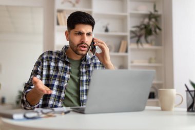 Kafası karışmış bir Arap cep telefonuyla dizüstü bilgisayar kullanarak konuşuyor.