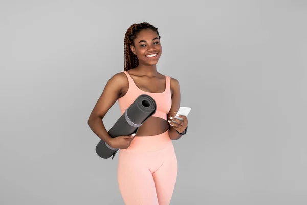 Atlético jovem mulher negra em sportswear segurando tapete de fitness e smartphone em fundo estúdio cinza — Fotografia de Stock