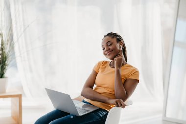 Genç siyahi kadın dizüstü bilgisayar kullanıyor, kulaklık takıyor, evde gözleri kapalı dinleniyor.